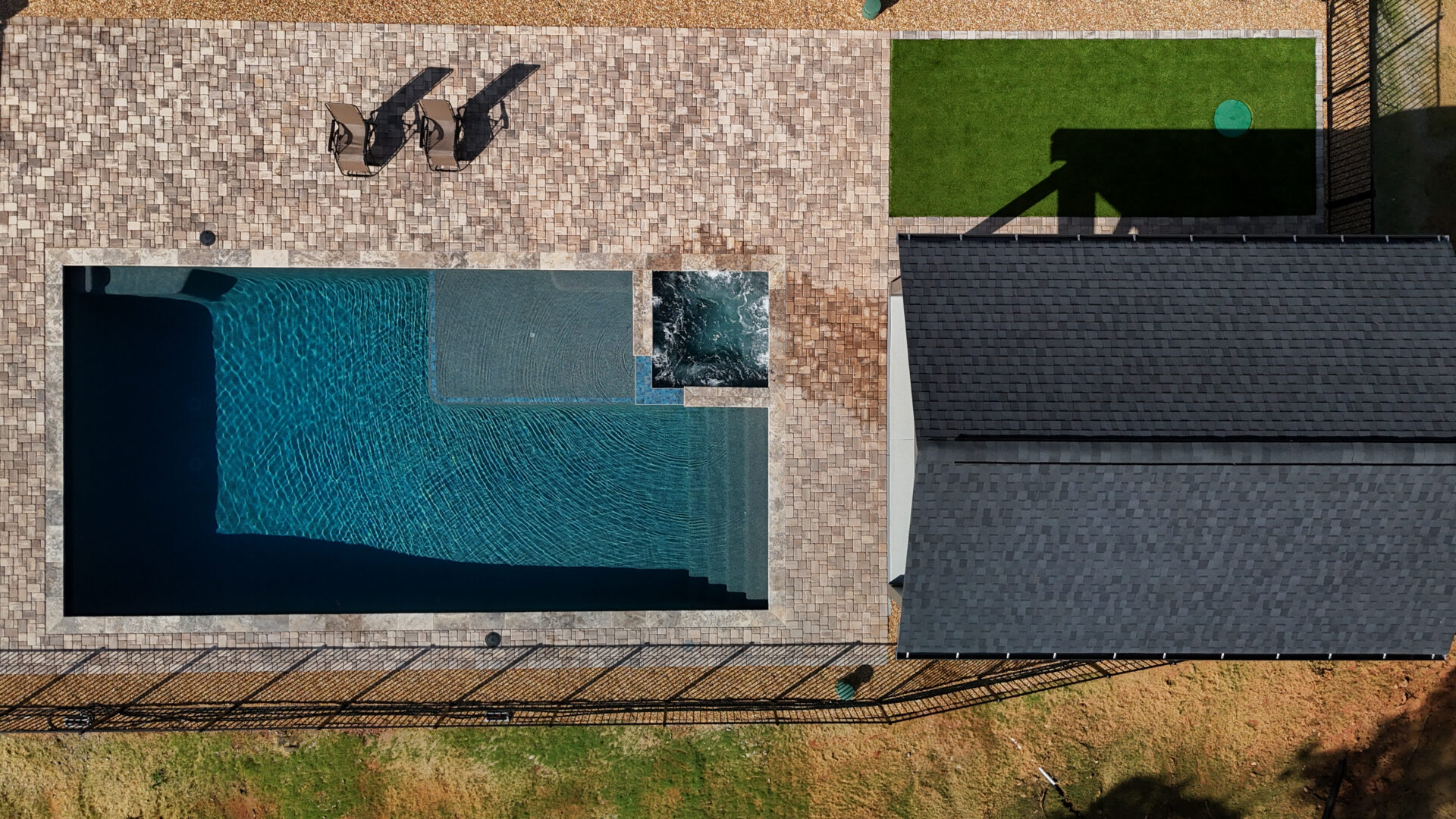 Aerial perspective of a backyard featuring a rectangular swimming pool, a small hot tub, patio chairs arranged on a paver deck, a vibrant green lawn area, and a dark-roofed structure, all surrounded by a fence.