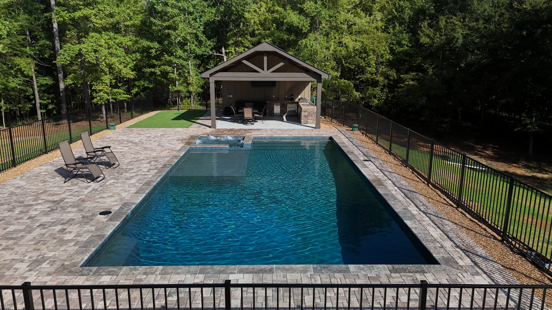 A rectangular swimming pool with a spa is surrounded by a stone patio and black fence. Two loungers sit on the left side. In the background, there’s a covered patio with tables and chairs nestled among tall trees.