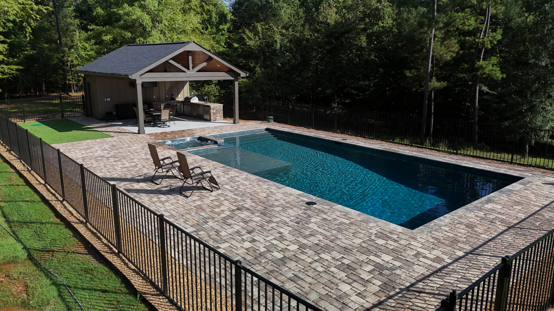 Rectangular pool with clear water, two lounge chairs on a brick patio, a small spa, a covered outdoor kitchen, and surrounding black metal fence. Trees and greenery line the background.