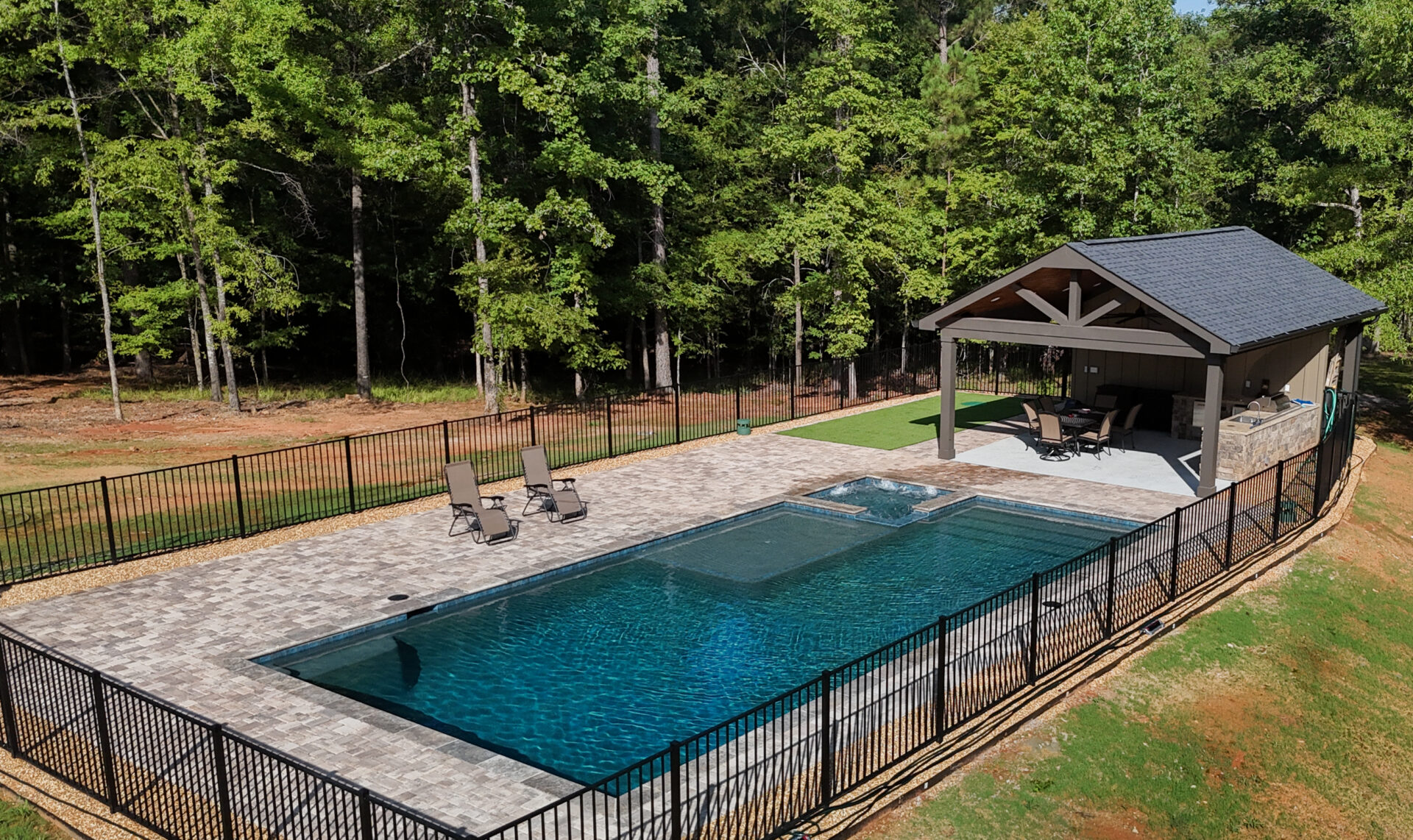 A fenced rectangular pool with lounge chairs arranged on paved ground, a nearby hot tub, and a covered patio furnished with tables and chairs, all surrounded by trees and lush greenery.