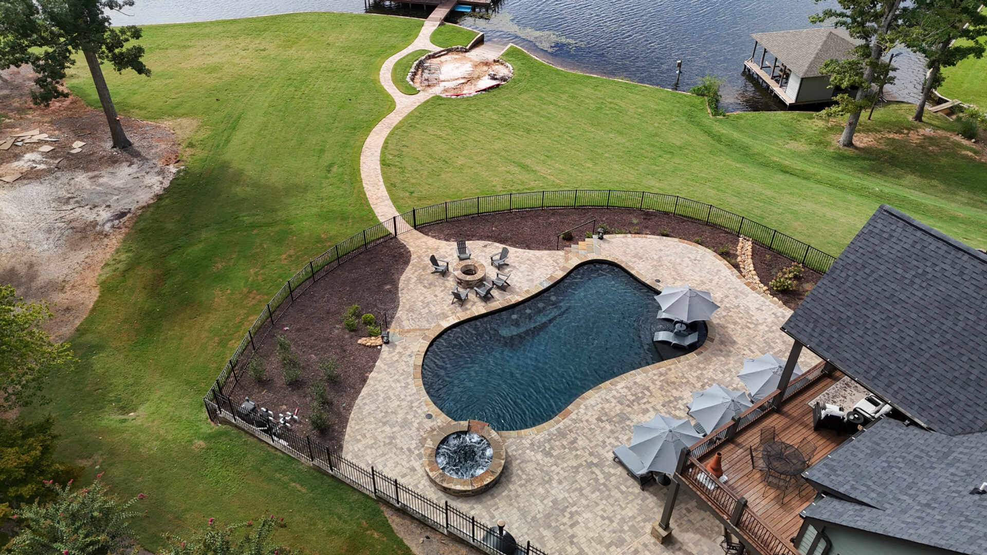 Aerial view of a backyard featuring a pool, hot tub, patio furniture, winding pathways, lush green lawn, and a dock extending to a lake. A waterfront gazebo and fire pit can also be seen.