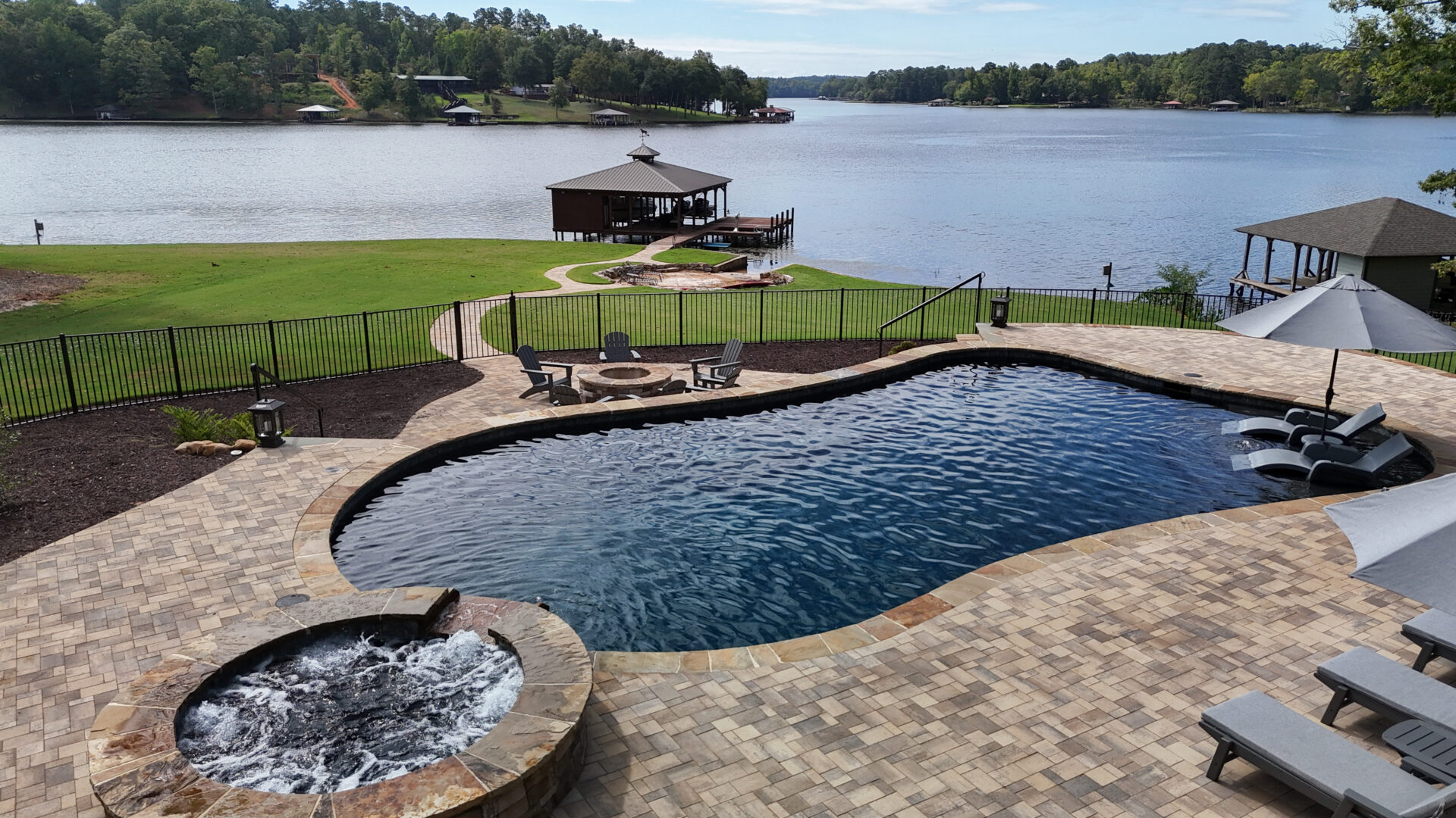Spacious backyard featuring a pool, hot tub, lounge chairs, and a fire pit, all overlooking a grassy lawn and a serene lake with boat docks and trees in the distance.