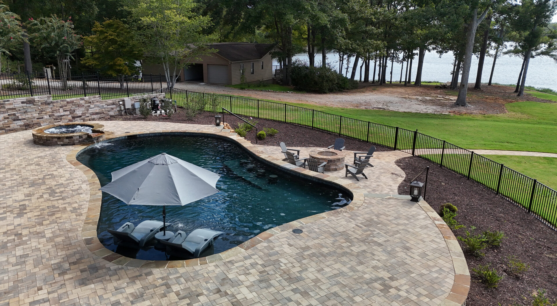 A spacious backyard features a curvy pool, lounge chairs shaded by an umbrella, a hot tub, a fire pit surrounded by chairs, lush trees, a garage, and scenic views of a lake in the background.