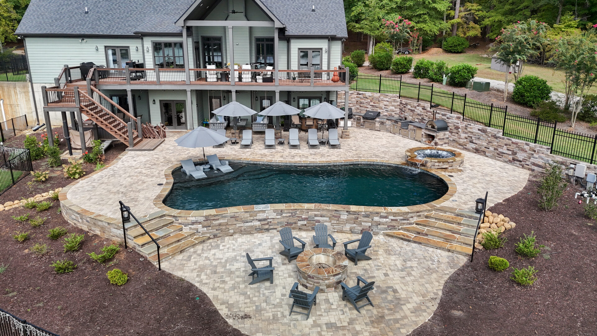 A spacious backyard showcases a stone patio with a swimming pool, several lounge chairs and umbrellas, a hot tub, a fire pit encircled by seating, and lush landscaping around a contemporary two-story home.