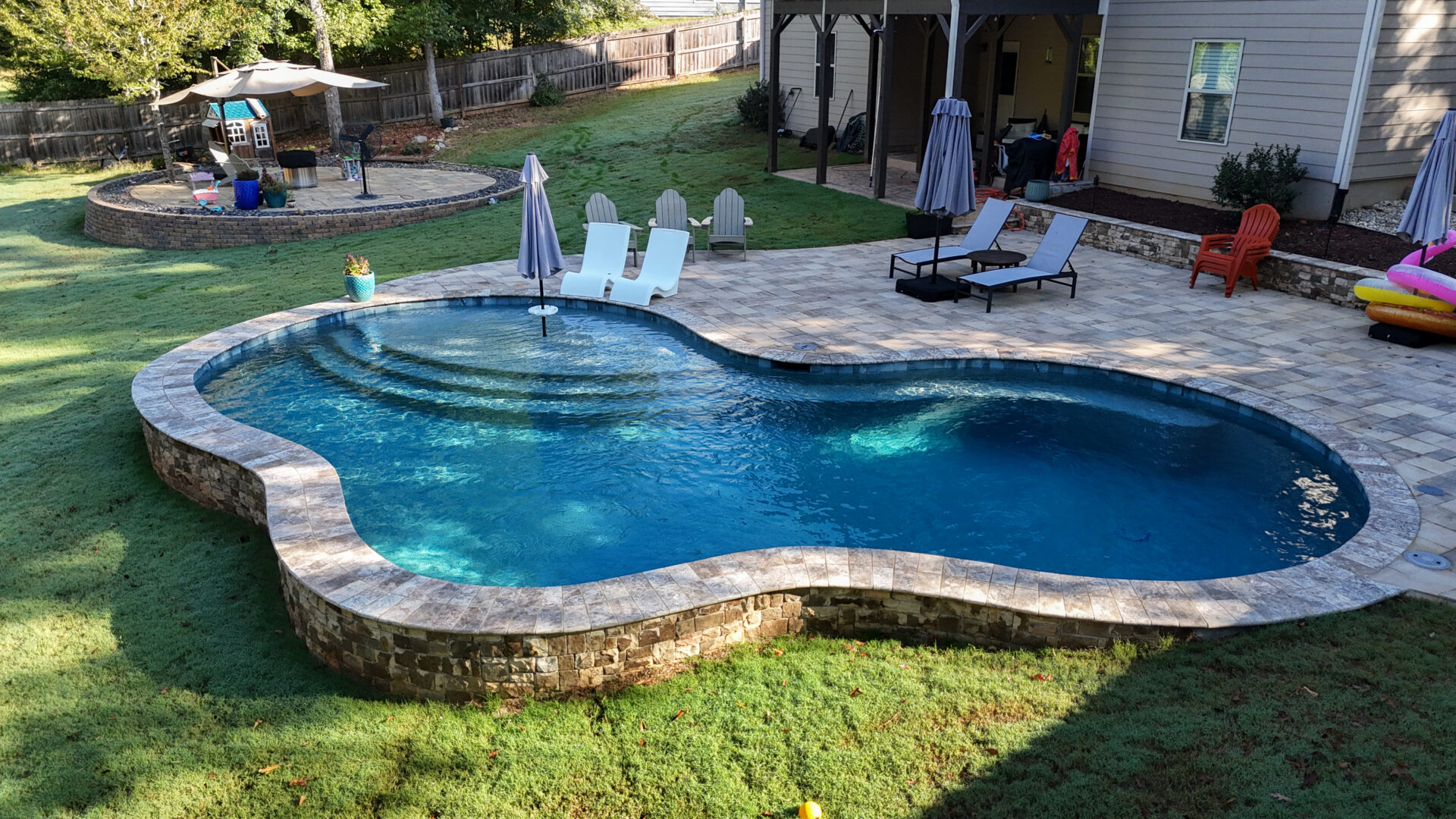 A backyard featuring a freeform swimming pool, lounge chairs, umbrellas, a patio area, and a play area, all surrounded by grass and a wooden fence.