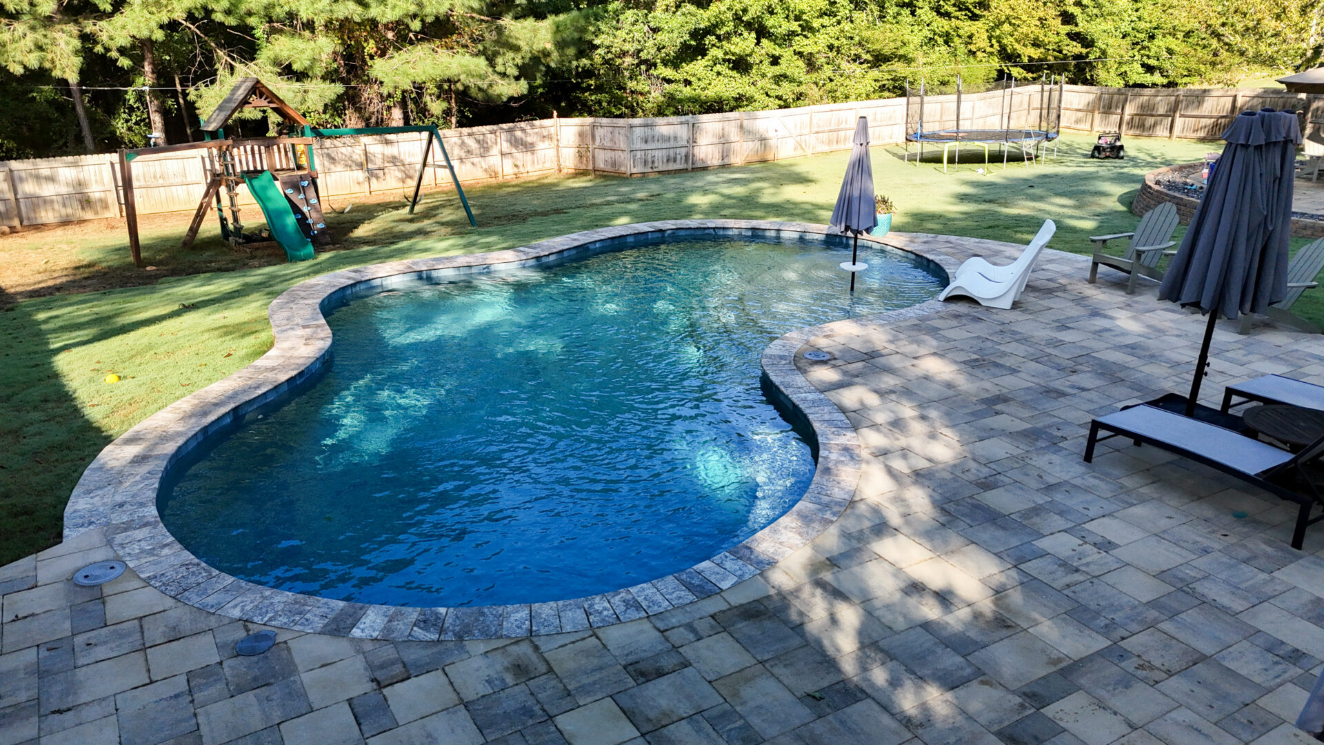 A backyard featuring a curvy pool, two lounge chairs with umbrellas, a wooden playset including a slide, a trampoline, and a fenced grassy area bordered by trees in the background.