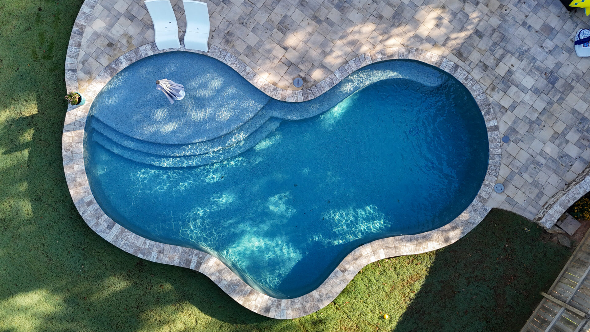 Aerial view of a kidney-shaped backyard swimming pool bordered by stone tiles and green grass, with two lounge chairs placed nearby.