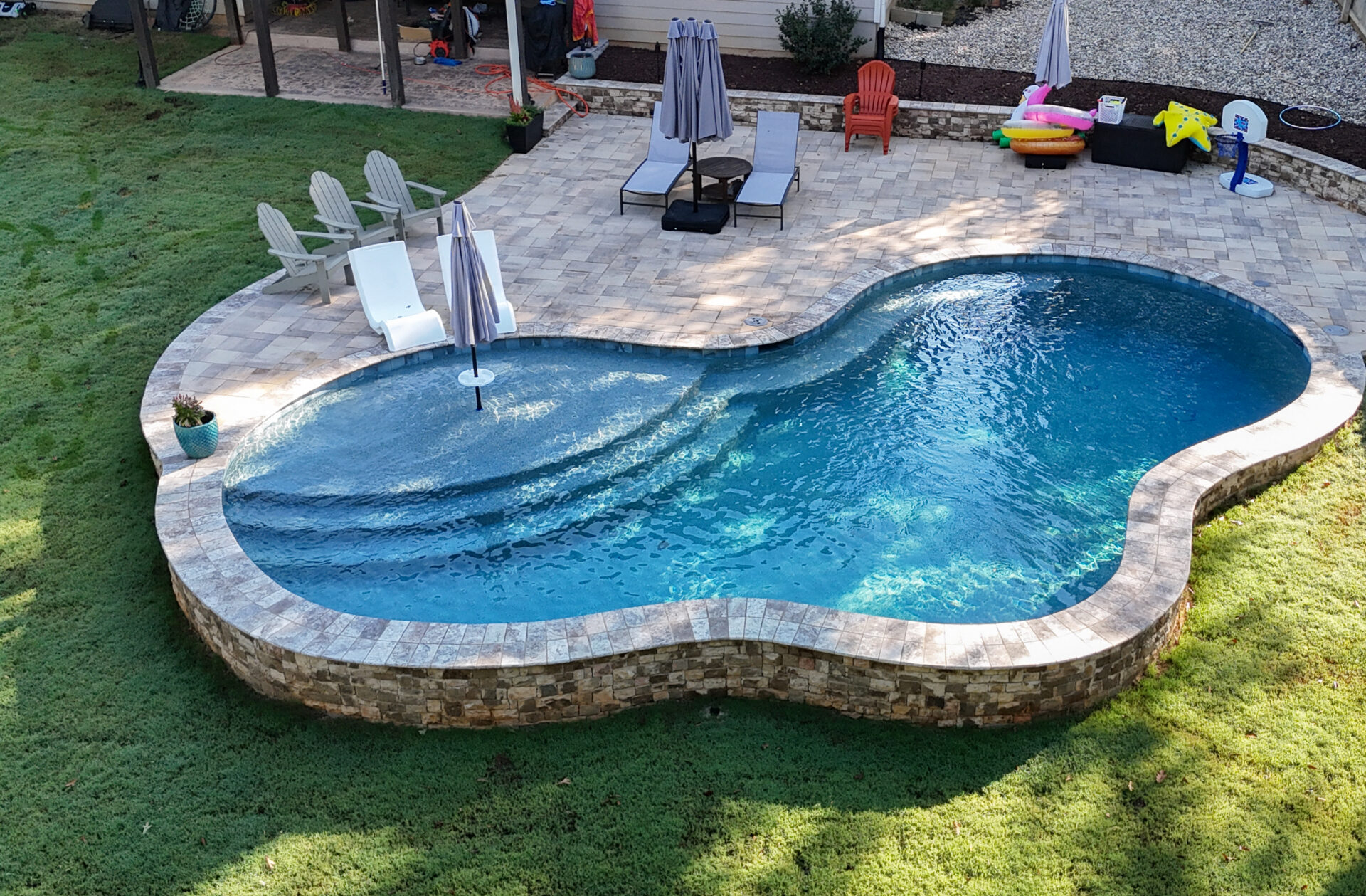 Backyard kidney-shaped pool with a stone border, surrounded by a patio, lounge chairs, umbrellas, a small table, and kids’ pool toys on the side. The area is bordered by grass.