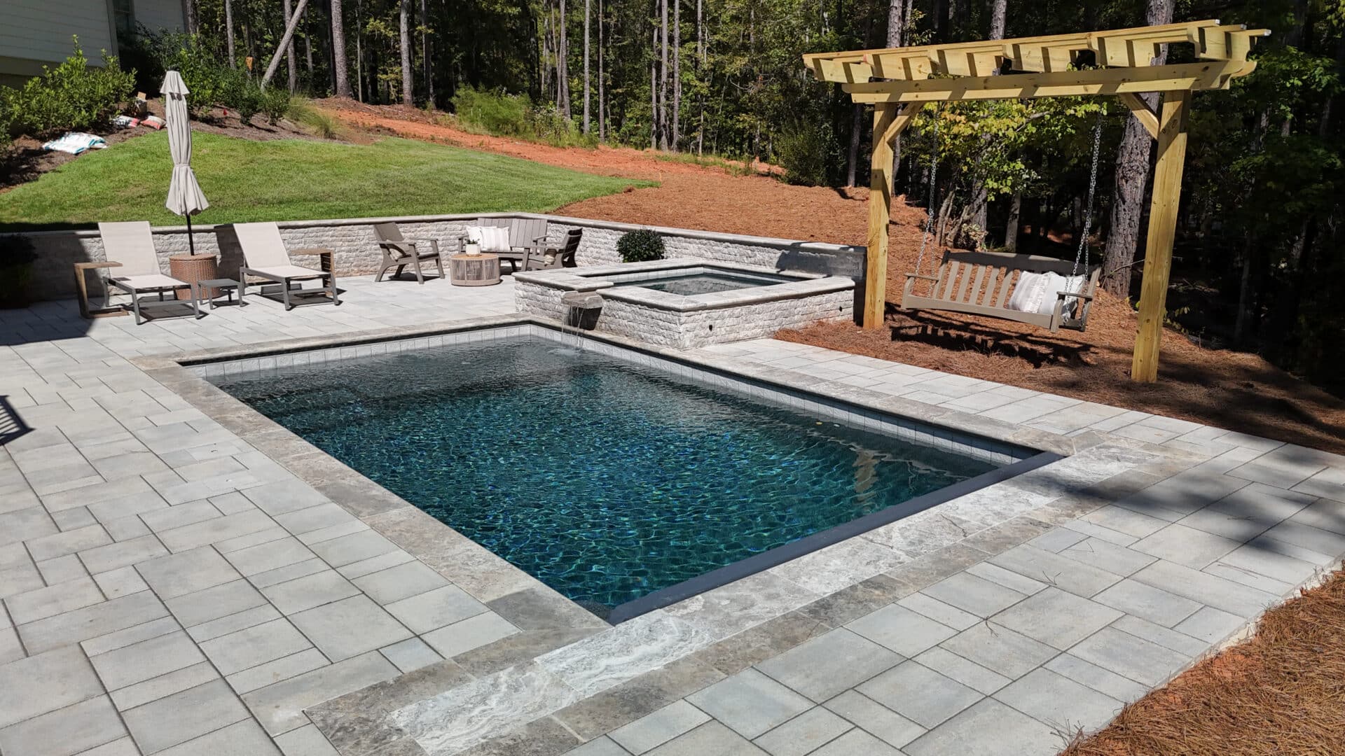 A backyard featuring a rectangular pool, hot tub, pergola swing, patio chairs, an umbrella, and a fire pit, all surrounded by trees and paved walkways.