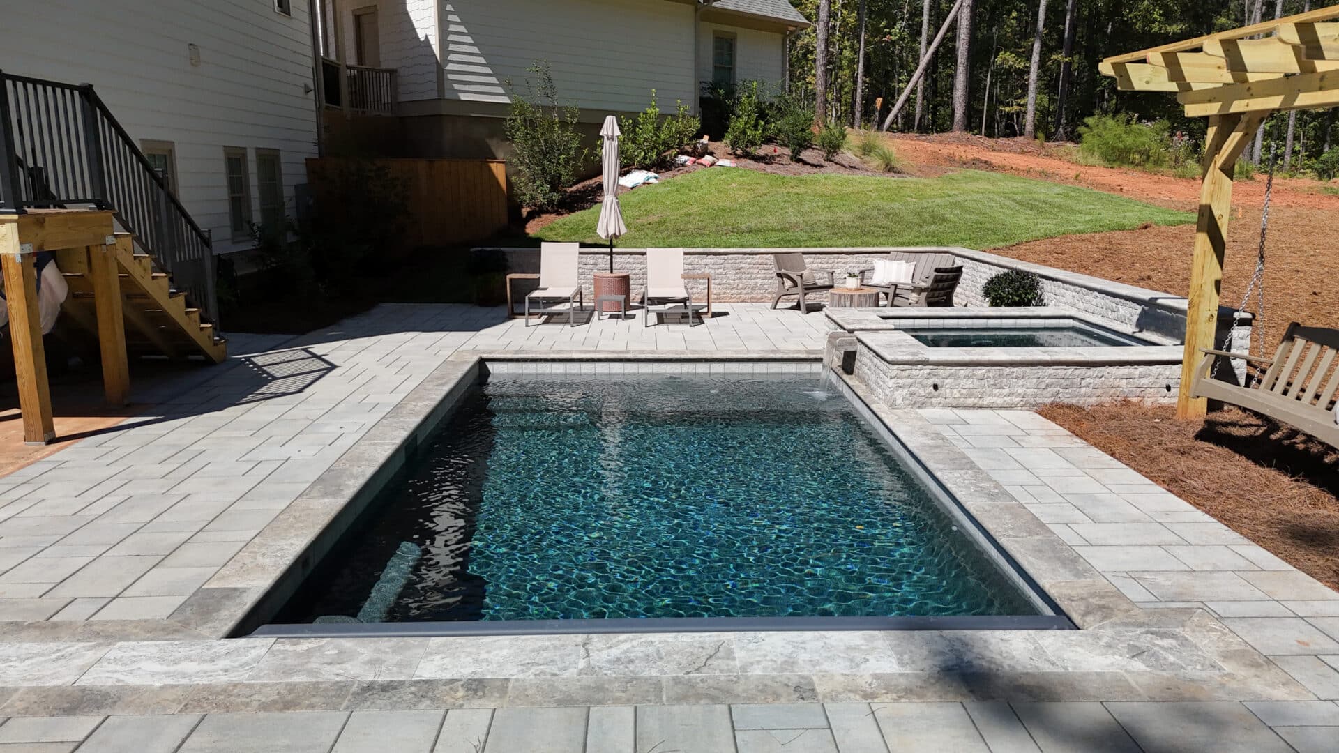 A rectangular backyard pool with clear water, bordered by stone pavers. Nearby, lounge chairs and umbrellas invite relaxation, while a raised hot tub and a landscaped yard complete the background scene.