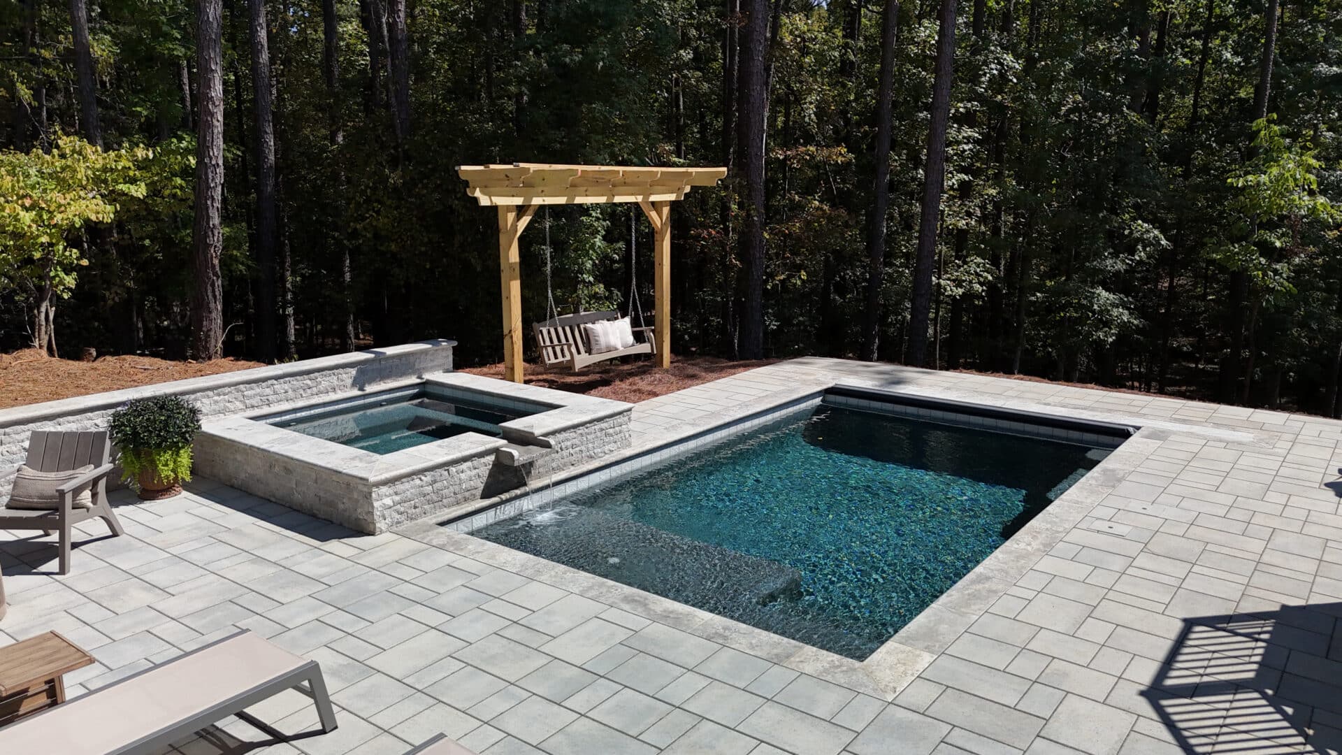 A backyard featuring a rectangular pool connected to a square hot tub, with patio seating nearby and a wooden pergola sheltering a swing. Tall pine trees in the background create a shaded, forested atmosphere.