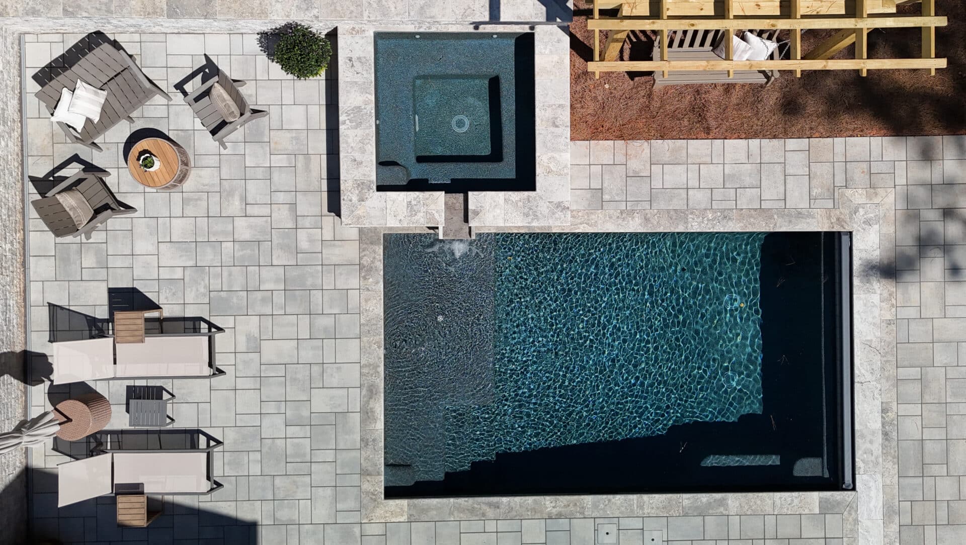 Aerial view of a backyard featuring a rectangular pool, square hot tub, lounge chairs, a small table, a pergola, and a paver stone patio. Shadows and greenery surround the area.