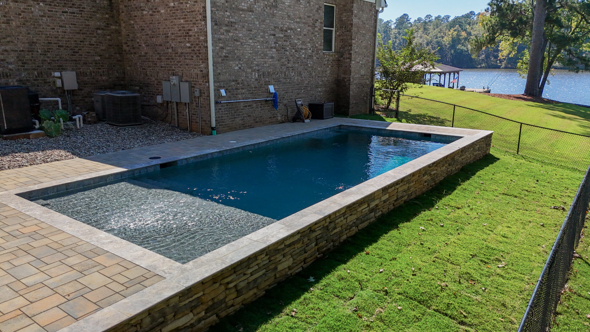 An elevated rectangular swimming pool with a shallow entry step situated in a backyard next to a brick house and grass lawn, with a lake and dock in the background.