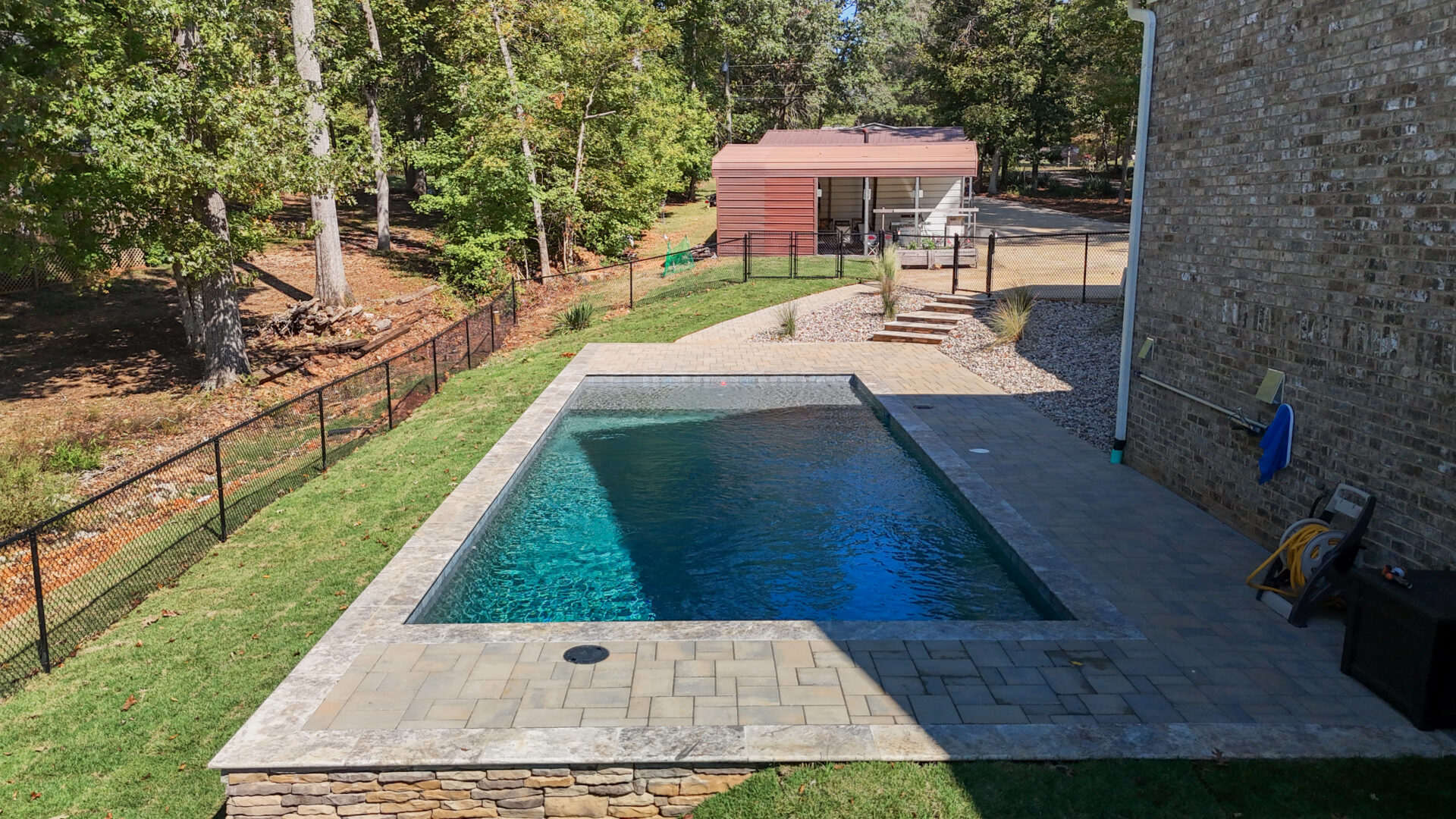 Rectangular swimming pool with a stone patio in a backyard with a black chain-link fence and a small shed in the background.