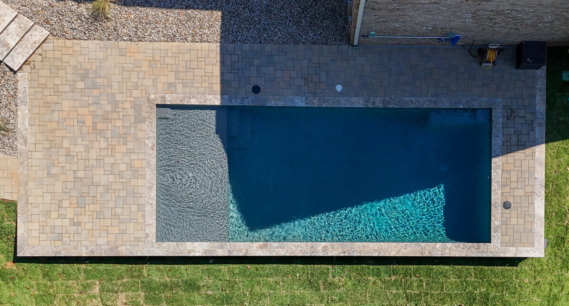 Aerial view of a rectangular swimming pool with a shallow tanning ledge, surrounded by a paved patio and grass.