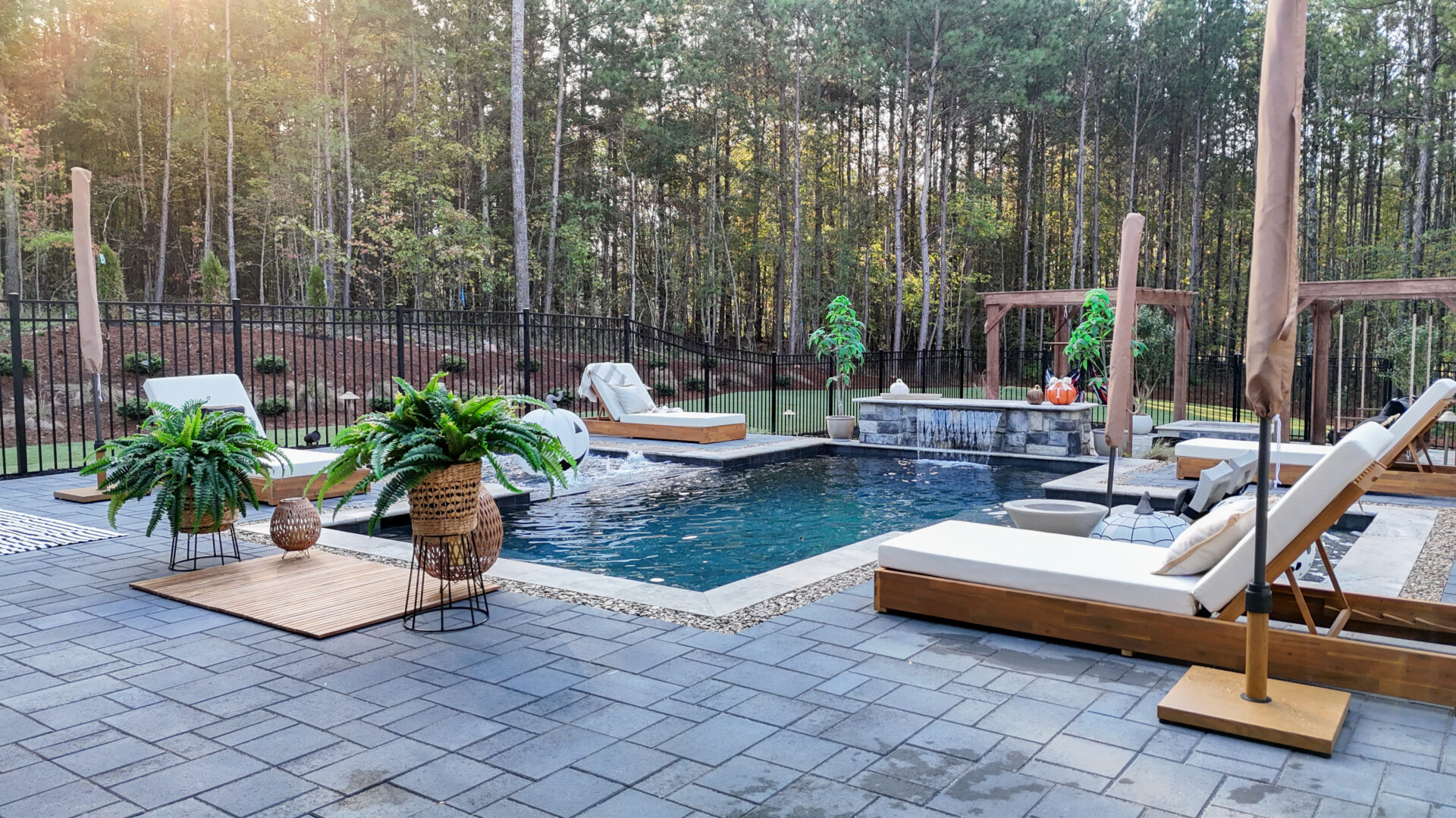 A swimming pool surrounded by a patio with multiple lounge chairs, potted ferns, and umbrellas, bordered by a fence and a forest.