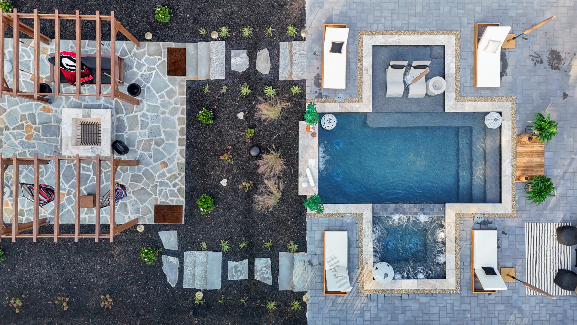 Aerial view of a backyard featuring a patio with a wooden pergola and fire pit on one side, and a swimming pool and hot tub surrounded by lounge chairs on the other.