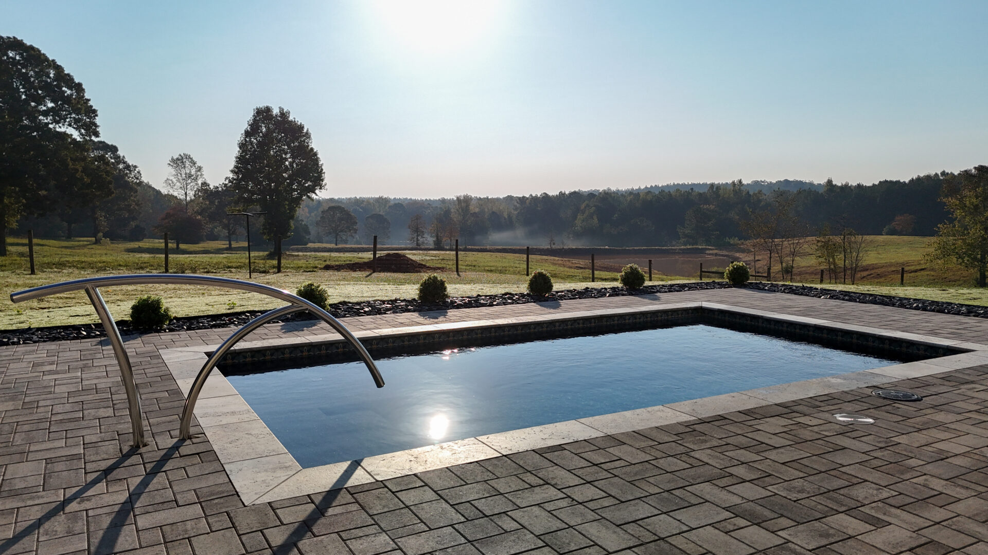 A rectangular backyard swimming pool with a metal handrail, surrounded by a paved patio, overlooking a field and forest at sunrise.