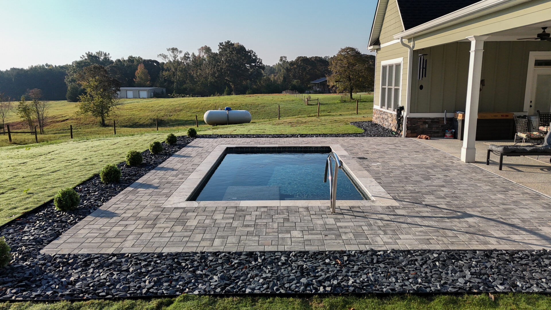 A rectangular inground pool set into a stone paver patio beside a house, overlooking a grassy field.