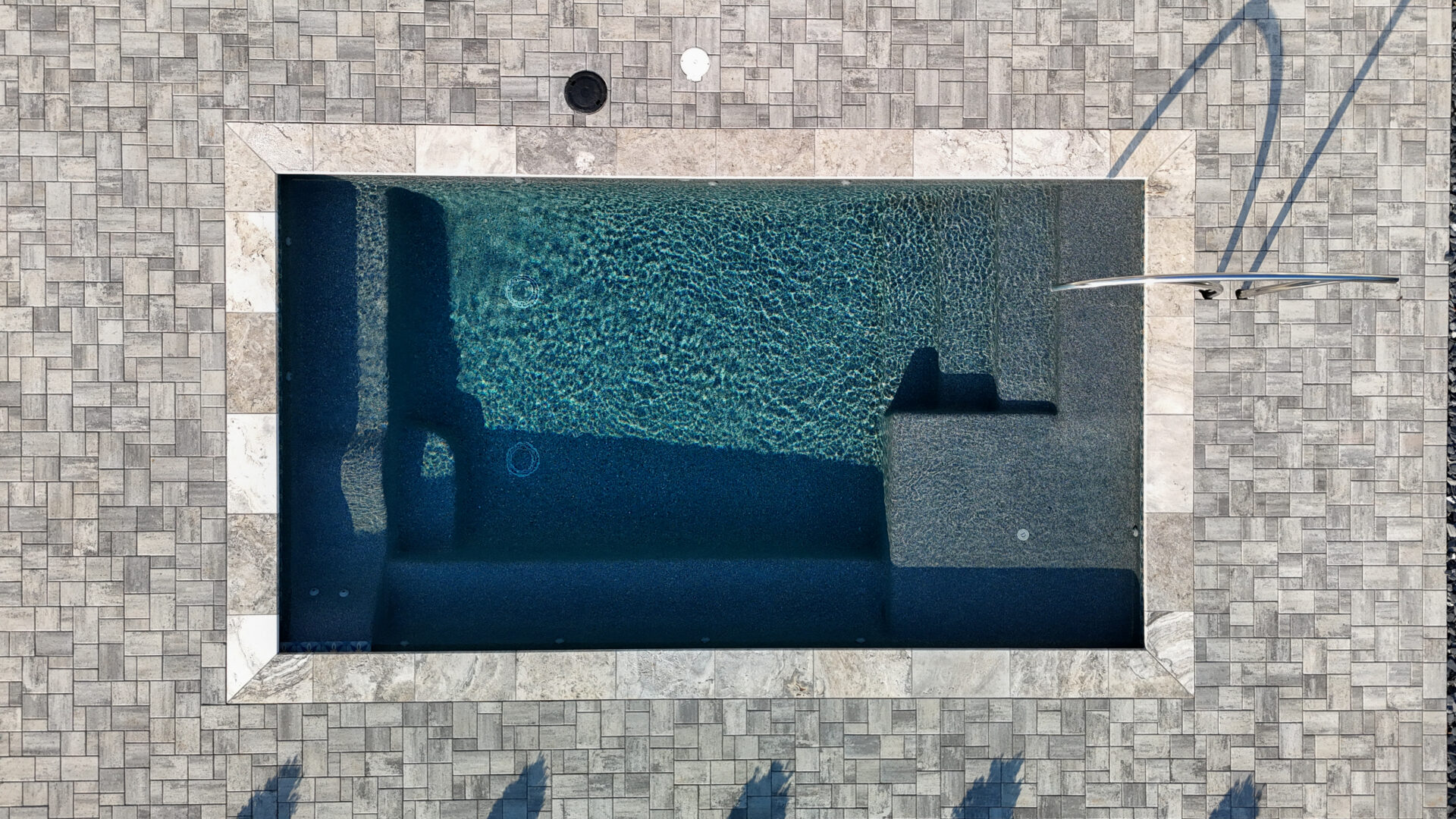 Aerial view of a rectangular swimming pool with integrated steps surrounded by a stone paver patio.