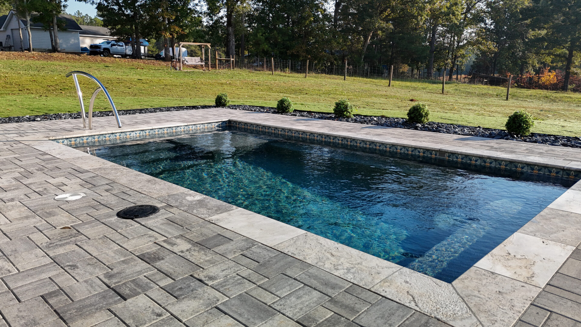 Rectangular swimming pool with dark blue water and a gray paver patio surround, featuring a metal handrail and decorative tile border.