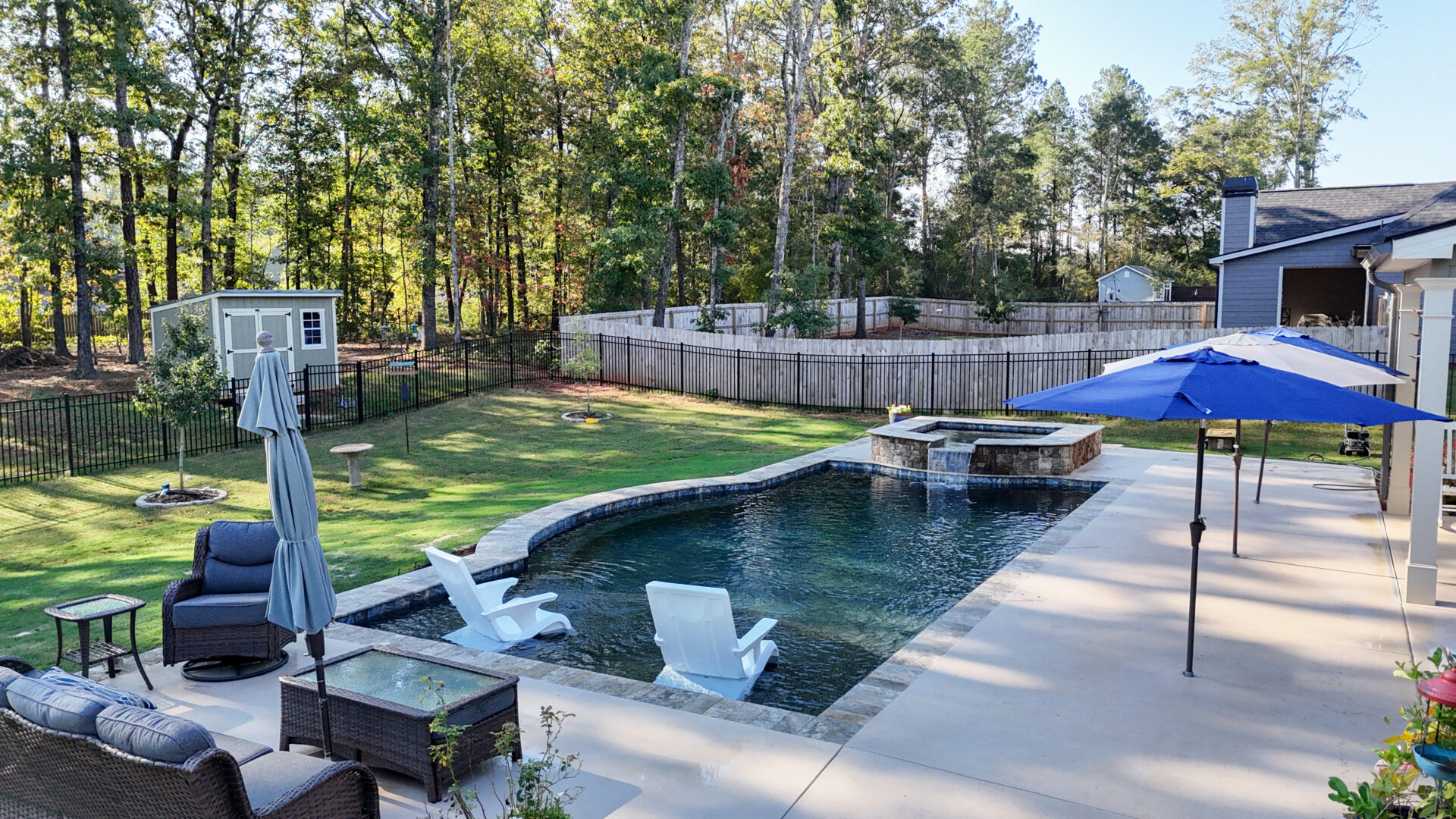 A backyard with an in-ground pool featuring a stone spillway, two white lounge chairs inside the shallow end, a patio with a table and chairs under blue umbrellas, and a fenced lawn with a storage shed.