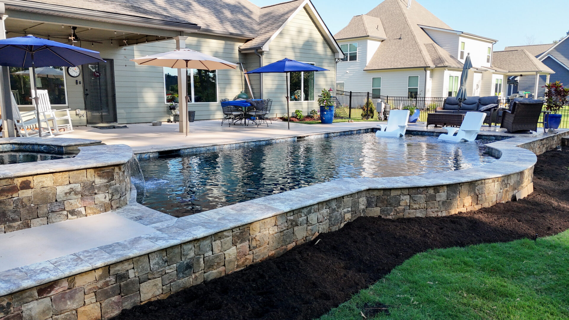 An elevated backyard swimming pool with a stone wall exterior, featuring two lounge chairs in the water and a patio area with umbrellas and furniture in the background.