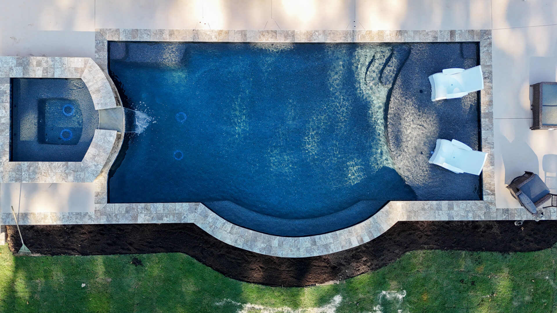 Aerial view of a rectangular swimming pool with a connected circular spa, featuring two white lounge chairs in the shallow tanning ledge.