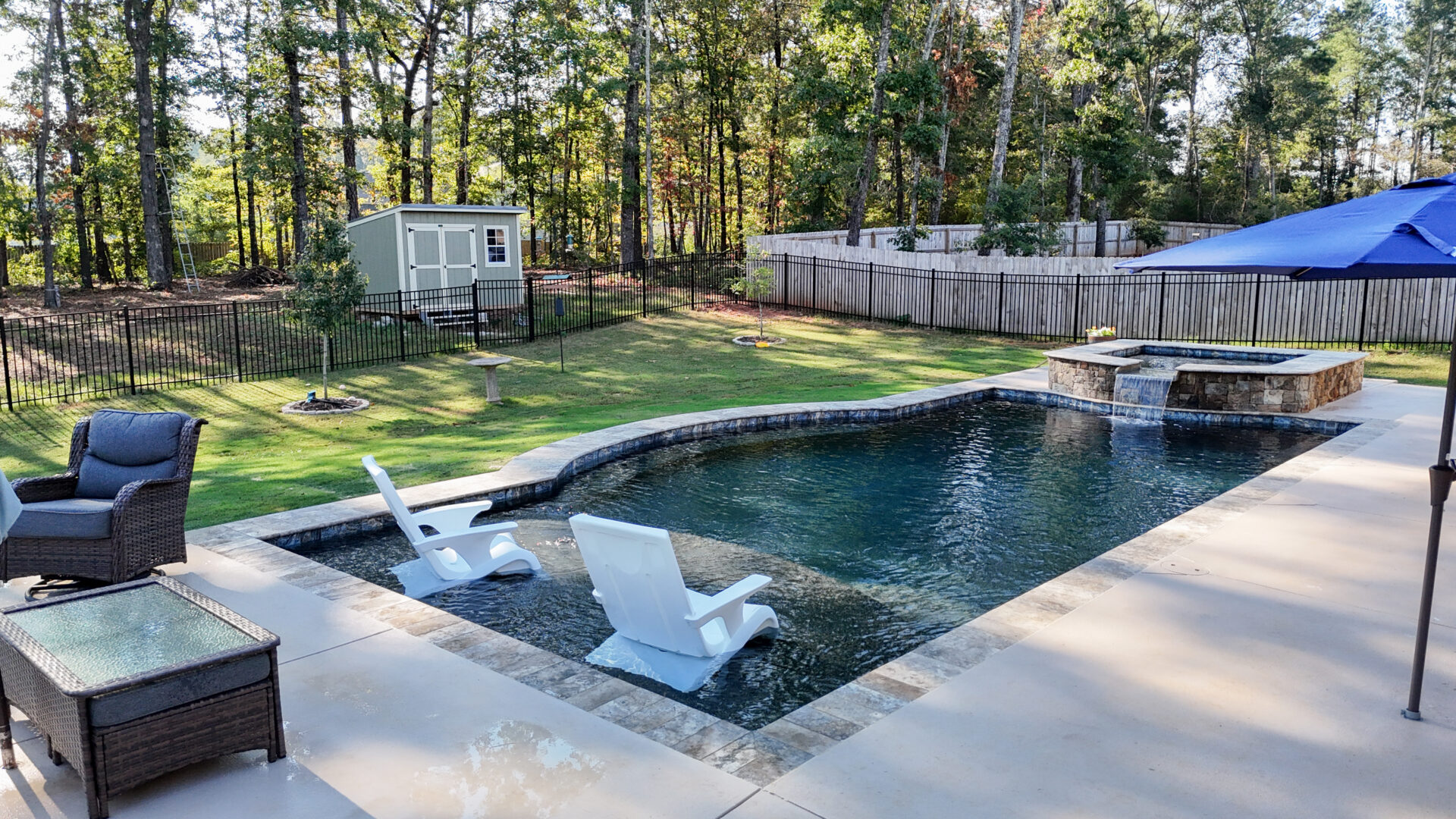 Two white lounge chairs sit in the shallow end of a swimming pool with a stone waterfall feature, next to a patio with wicker furniture.